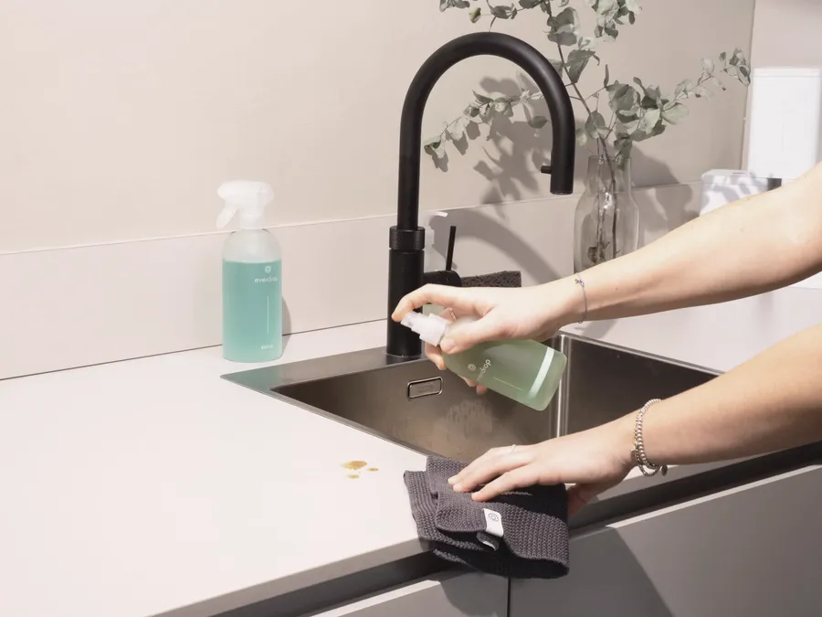 A hand spraying surface cleaner onto a cloth beside a kitchen sink